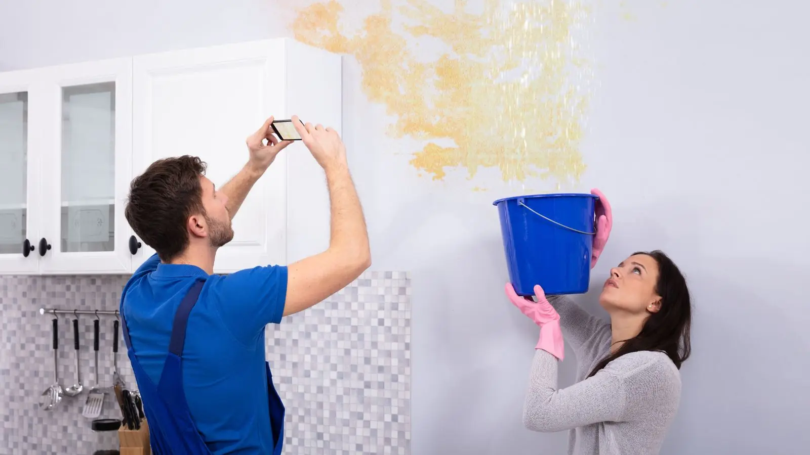 Homeowner photographing a water stain on the ceiling while another person holds a bucket